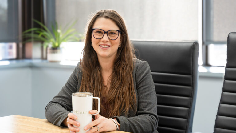 Christina Morgan, Partner at Welch LLP Cornwall, sits at a desk holding a Welch LLP tumbler and smiles at the camera