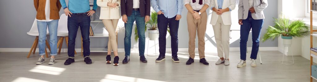 Unidentifiable people standing in a row wearing casual attire at a workplace. Clothing features jeans, sweaters, t-shirts, sneakers, loafers, and slacks.