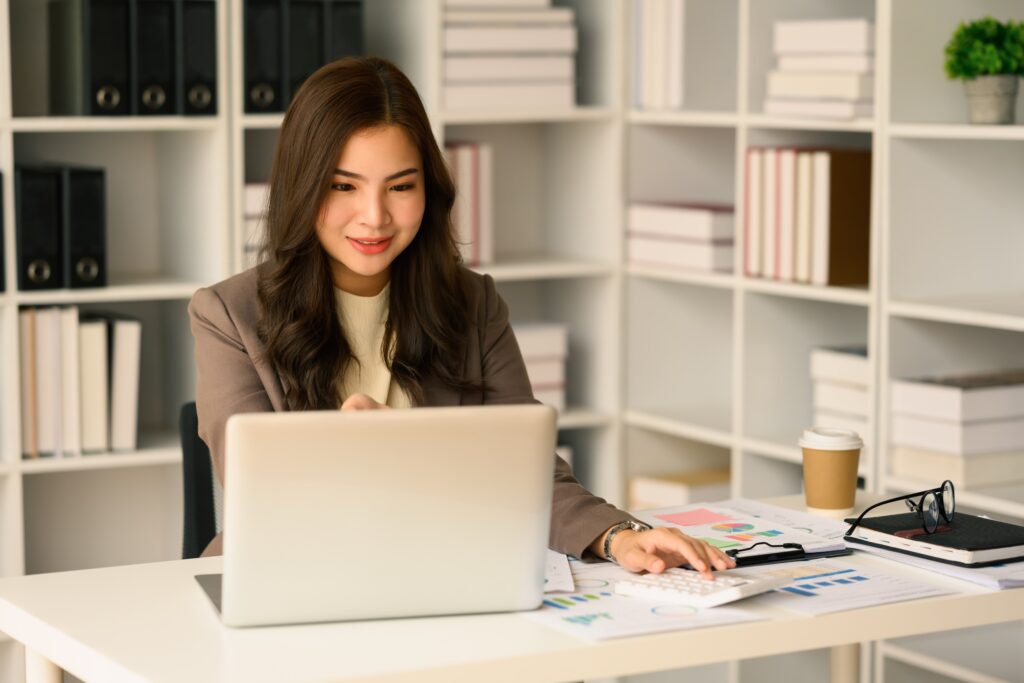 A young woman sits a desk working with papers, calculator, and a computer